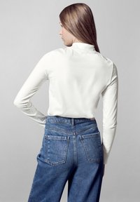 Weißes Langarmoberteil mit hohem Halsabschluss, kombiniert mit hochgeschnittenen blauen Denim-Jeans mit klassischem Fünf-Taschen-Design.
