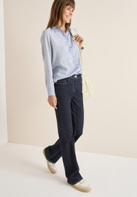 Camicia azzurro chiaro a righe, jeans in denim scuro e sneakers bianche con suole beige. Il modello porta una borsa a tracolla color crema.
