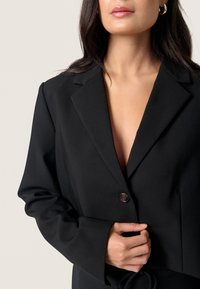 Czarna marynarka z klapami, zapinana na jeden guzik, z szerokimi rękawami. Wykonana z gładkiego materiału, skrojona w dopasowanym fasonie.