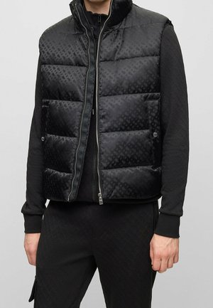 Man wearing a black quilted puffer vest with front zipper over a black long-sleeve shirt and black pants against gray background.