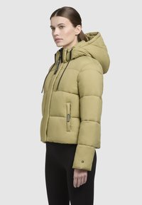 Jachetă puffer khaki cu glugă, având un design matlasat, sfori negre și accente cu fermoar. Include mâneci lungi și buzunare laterale.