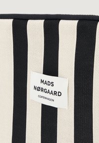 Ριγέ τσάντα σε μαύρο και κρεμ, κατασκευασμένη από υλικό με υφή. Διαθέτει ετικέτα με τη γραφή "MADS NØRGAARD COPENHAGEN" σε έντονο μαύρο κείμενο. Κλείσιμο με φερμουάρ.
