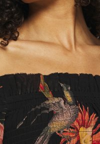 Spalla e collo di una donna che indossa un top nero senza spalle con una stampa colorata di uccelli e fiori. Capelli ricci scuri visibili.