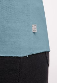 Turquoise gebreid top met een textuurafwerking, voorzien van een klein stoffen label met een hondontwerp, afgewerkt met een ruwe rand. Gepaarde met donkere stof.