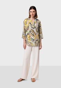 Tunic multicolore con intricati motivi paisley in giallo, verde e marrone; presenta un colletto a V e maniche a tre quarti. Abbinata a pantaloni bianchi a gamba larga.
