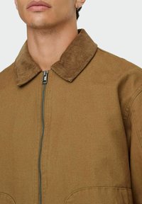 Pogled od blizu na osebo, ki nosi rjav zip-up jakno s kordurojnim ovratnikom in uhani na levem ušesu.