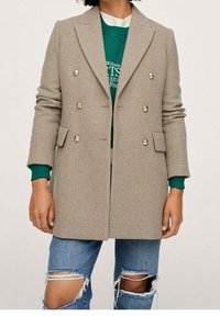 Cappotto doppiopetto grigio chiaro con bottoni argento, design sartoriale e tessuto misto lana. Indossato sopra una felpa verde e jeans blu strappati.