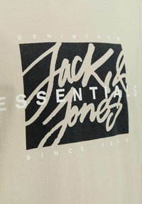 Helles beiges T-Shirt aus Baumwolle, mit einem großen schwarzen Grafikdesign und weißem Text, der "Jack & Jones ESSENTIALS" und "DENIMCREW" liest.