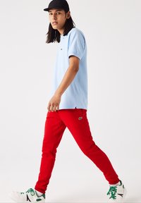 Lyseblå polo skjorte, røde bukser med logo, sort kasket, hvide sneakers med grønne detaljer, gående pose. Enkel design og livlige farver.