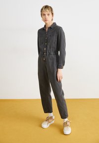 Salopette in denim grigio scuro con colletto, chiusura a bottoni, due tasche sul petto e vita elastica. Abbinata a sneakers con piattaforma caratterizzate da dettagli beige e bianchi.