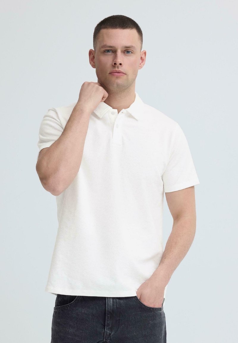 Homme aux cheveux courts portant un polo blanc et un jean foncé, debout devant un fond clair uni, main près du col et de la poche.