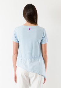 Lysblå kortærmet top med asymmetrisk kant, blød tekstur og et pink mærke ved nakken. Enkel, afslappet design.