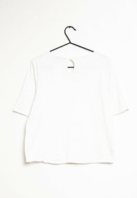 Top bianco a maniche corte con un motivo a diamante testurizzato e una fessura a chiave sulla schiena, appeso a un gancio nero su uno sfondo chiaro.