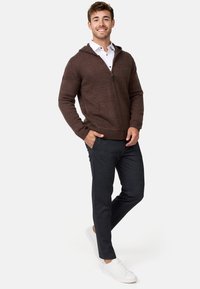 Brauner Reißverschluss-Hoodie mit strukturiertem Strick, Kängurutaschen und silbernem Reißverschluss, kombiniert mit dunklen Hosen und weißen Sneakers.