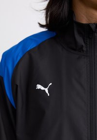 Crna jakna s plavim akcentima, koja ima visoki ovratnik i zatvarač. Puma logo je istaknuto prikazan u bijeloj boji na području prsiju.