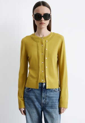 Vrouw die een mosterdgele cardigan met knopen draagt over een bijpassende top en blauwe spijkerbroek, met grote zwarte zonnebril, staand tegen een witte achtergrond.