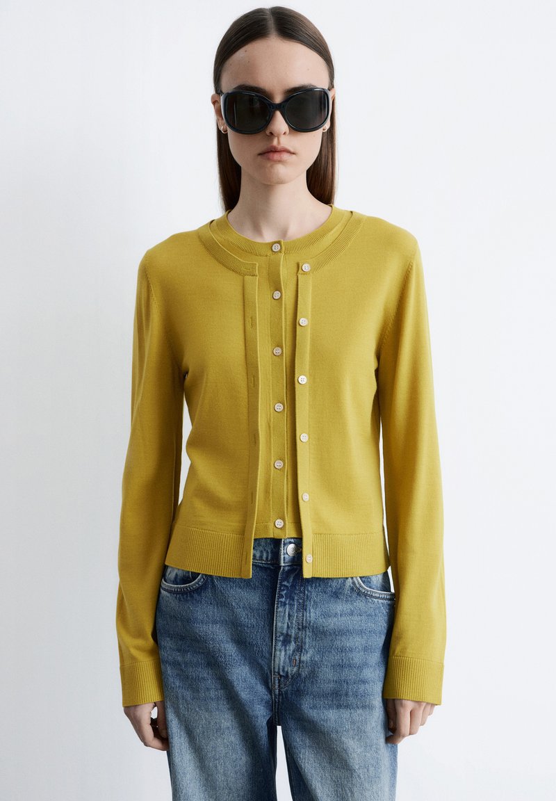 Femme portant un cardigan boutonné jaune moutarde sur un haut assorti et un jean en denim bleu, avec de grandes lunettes de soleil noires, debout sur un fond blanc.