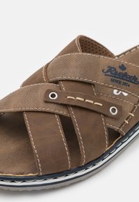 Braune Sandale mit kreuzgeknüpften Riemen, aus strukturiertem Material. Mit dekorativer Naht und einem blauen Logo am Riemen. Weiße Sohle als Akzent.