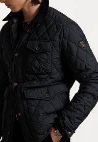 Czarna pikowana kurtka z kołnierzem z fartucha, wieloma kieszeniami, guzikami w kolorze złota oraz małą naszywką z logo na rękawie. Średnia tekstura.