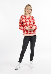 Pulover houndstooth roșu și roz cu mânici și margine inferioară cu nervuri. Asortat cu blugi negri și adidași albi. Modelul stă cu mâinile împreunate.