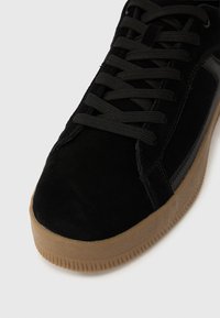 Baskets en daim noir avec lacets foncés et semelle en gomme marron, présentant des coutures détaillées et une semelle extérieure texturée sur fond blanc.