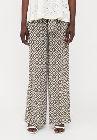 Pantalons larges imprimés en marron avec un motif en losange blanc, dotés d'une taille élastique avec cordon de serrage. Portés avec des sandales blanches.