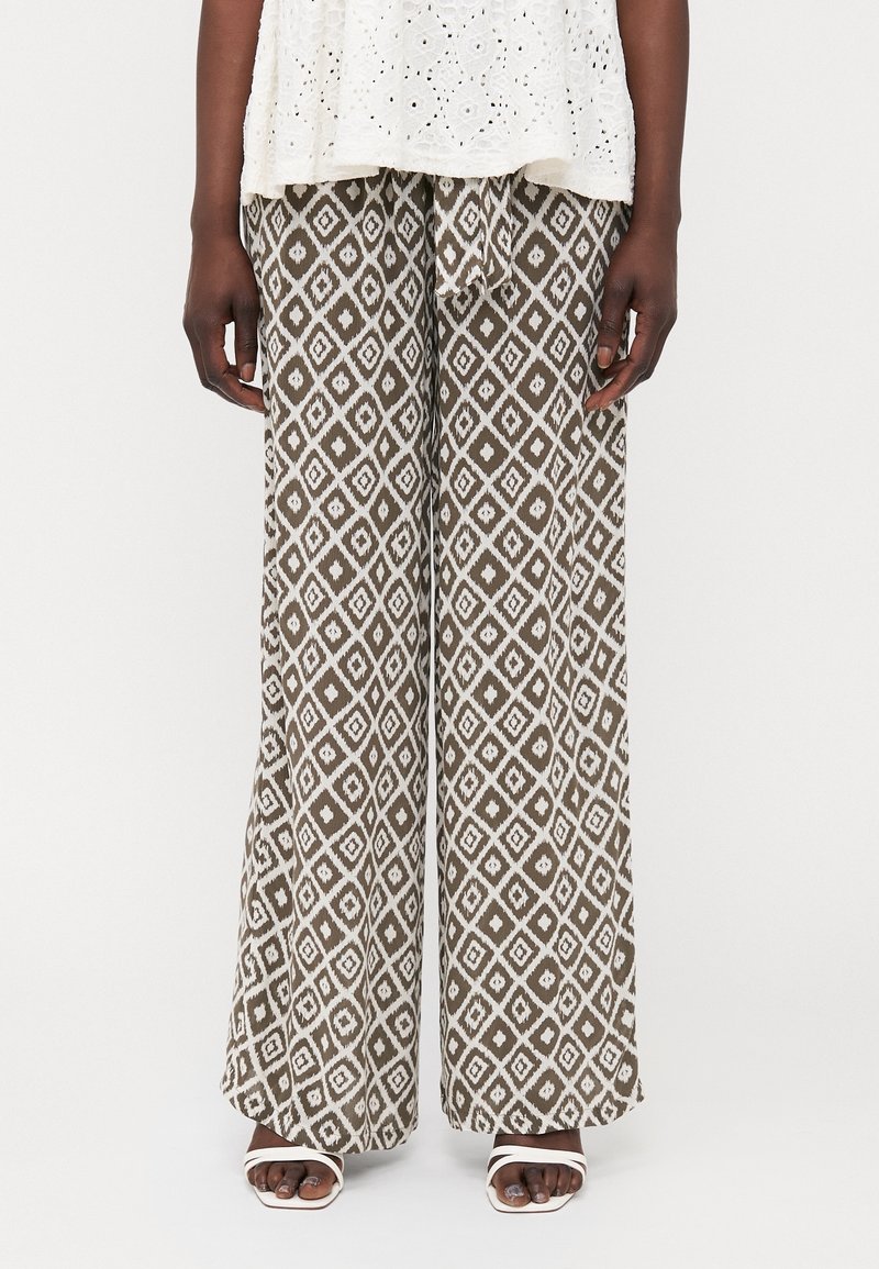 Pantalons larges imprimés en marron avec un motif en losange blanc, dotés d'une taille élastique avec cordon de serrage. Portés avec des sandales blanches.
