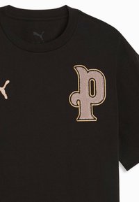 T-shirt noir Puma avec un écusson beige texturé en forme de "p" sur la poitrine droite et un petit logo Puma beige sur la poitrine gauche.