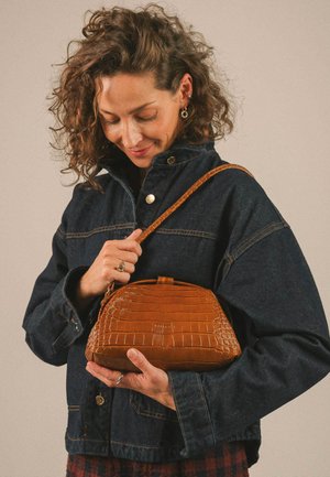 Femme aux cheveux bouclés portant une veste en jean foncé tenant un sac à main en cuir texturé marron avec une bandoulière, regardant vers le bas.