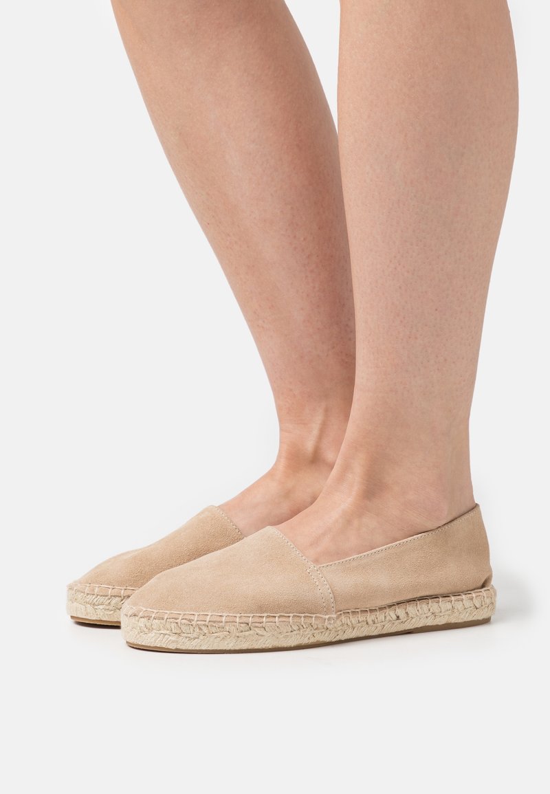 Pieds portant des espadrilles en daim beige avec des semelles en jute tressée, posés sur un fond clair uni.