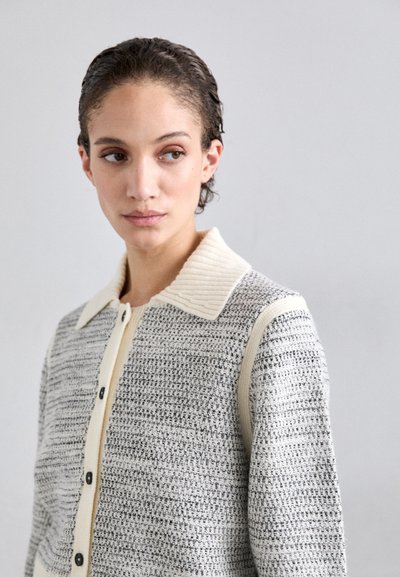 Strikket cardigan i grå og creme, med et struktureret mønster, en ribbet krave og knaplukning med kontrastfulde sorte knapper.