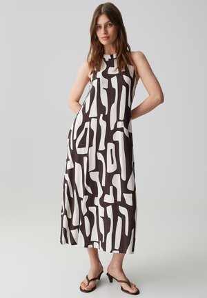 Frau trägt ein ärmelloses schwarz-weiß abstrakt gemustertes Midikleid mit braunen Sandalen mit Absatz, steht mit Händen in der Hüfte vor einem einfarbigen Hintergrund.