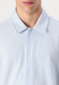 Polo de color azul claro, texturizado, con cuello y tres botones. Tejido suave con una superficie ligeramente borrosa.