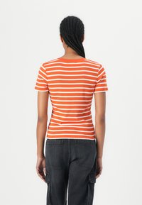 Oransje og hvitstripete T-skjorte med kort erme, tettsittende design, bakvinkel. Kombinert med svarte cargo-bukser med sidelommer.