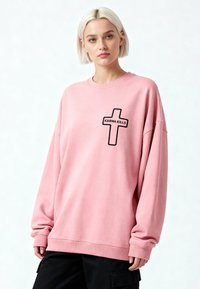 Jeune femme aux cheveux blonds courts portant un sweat-shirt rose oversize avec une croix noire et le texte "KARMA KILLS", associé à un pantalon noir.