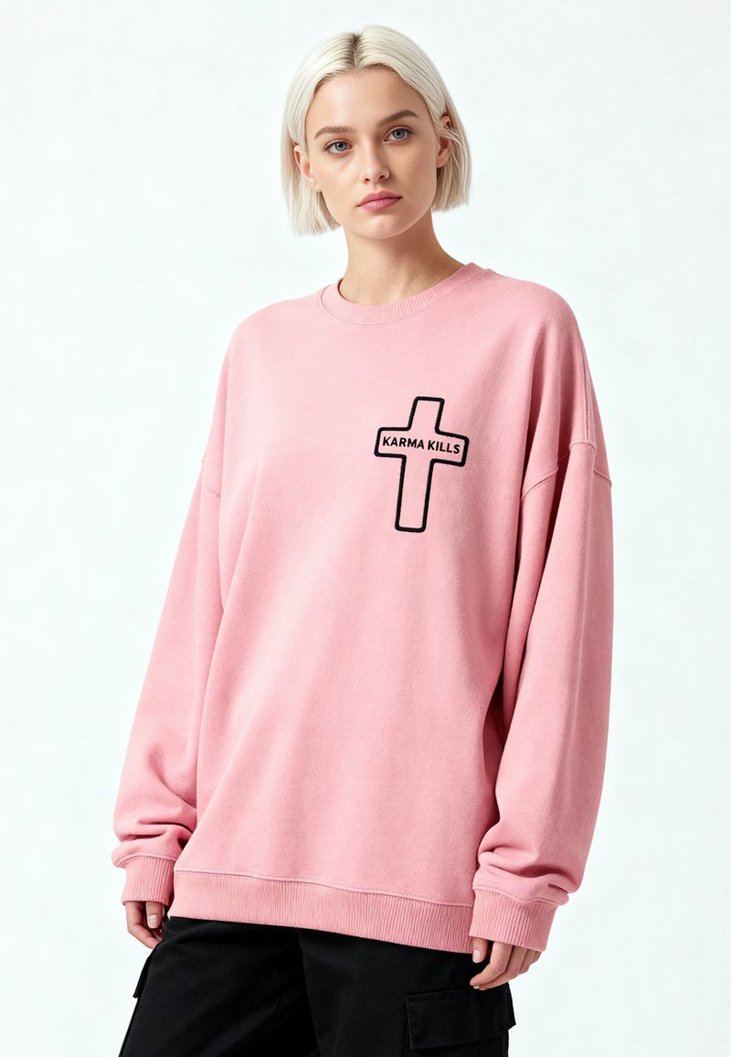 Jeune femme aux cheveux blonds courts portant un sweat-shirt rose oversize avec une croix noire et le texte "KARMA KILLS", associé à un pantalon noir.