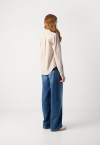 Camicia a righe in crema e beige, con orlo arrotondato e maniche lunghe, abbinata a pantaloni jeans blu larghi e sneakers bianche.