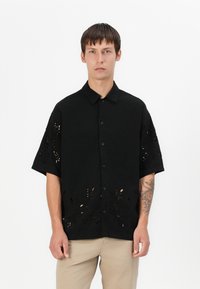 Μαύρο κοντομάνικο πουκάμισο με καπέλο κουμπιού, με κομμένα λουλουδάτα σχέδια στο στρίφωμα και στα μανίκια, κατασκευασμένο από απαλό, ελαφρώς υφασμάτινο ύφασμα.