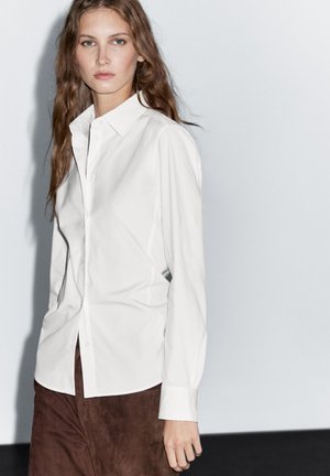 White long-sleeve button-up shirt, structured cotton with a smooth texture, paired with brown suede trousers. Simple collar and straight fit.