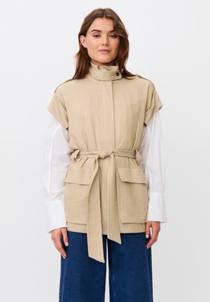 Donna con gilet beige con cintura sopra una camicia bianca a maniche lunghe e jeans blu, in piedi davanti a uno sfondo chiaro e uniforme.