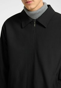 Chaqueta negra con cierre de cremallera, cuello con solapa y un suéter de cuello alto acanalado en gris claro debajo. Tela suave, diseño minimalista, sin patrones visibles.