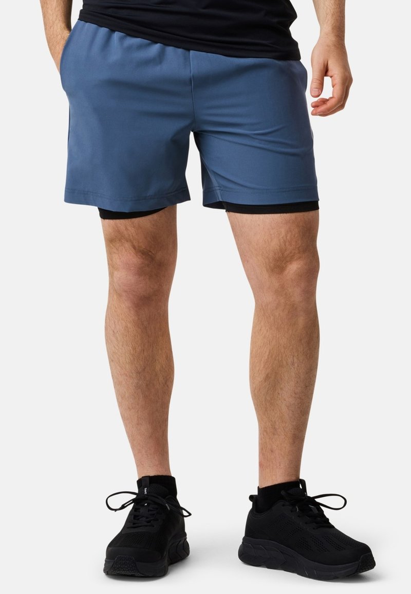 Mäns blåa sportshorts med svart innerfoder, elastisk midja, slät textur och knälång skärning. Bärs med svarta sneakers.