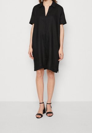 Frau trägt ein locker sitzendes, schwarzes, kurzärmliges Kleid mit Kragen und Frontschlitz, kombiniert mit schwarzen Sandalen mit Knöchelriemen und Blockabsatz.