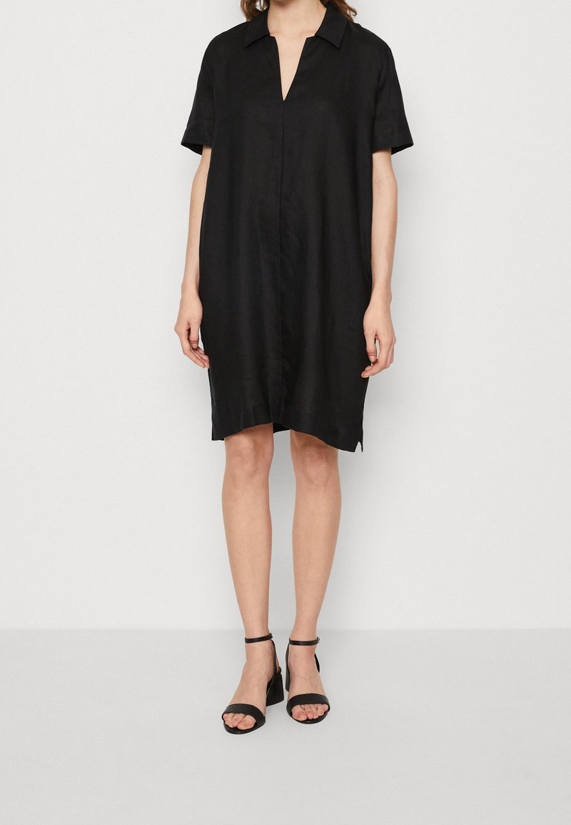 Femme portant une robe noire ample à manches courtes avec col et fente à l'avant, associée à des sandales à talons blocs avec bride à la cheville.