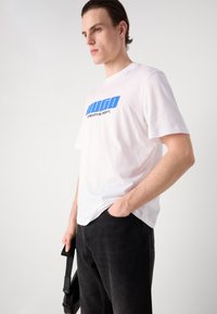 Camiseta de algodón blanca con estampado gráfico azul "HUGO CREATIVE DEPT.", de manga corta, corte relajado, combinada con pantalones negros texturizados.