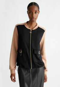 Giacca bomber color block nera e beige con zip frontale, due tasche frontali e maniche a contrasto. Realizzata in un tessuto liscio.