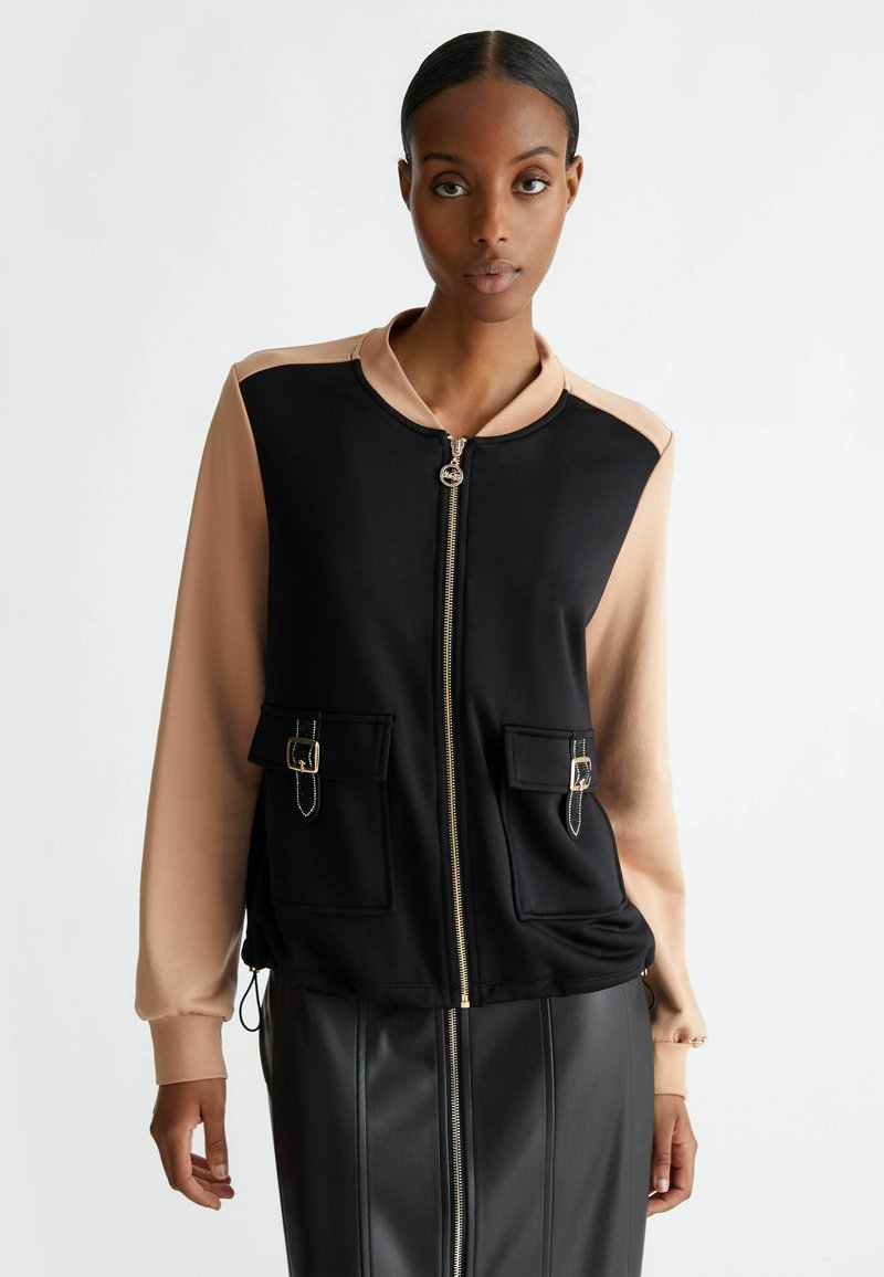 Giacca bomber color block nera e beige con zip frontale, due tasche frontali e maniche a contrasto. Realizzata in un tessuto liscio.