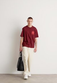 Burgundy cotton T-shirt with front logo, paired with cream wide-leg pants and white sneakers. Model carries a black bag. Plain background.