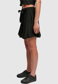 Schwarzer, knielanger Rock mit taillierter Taille und Bindegürtel, versehen mit Falten und einem ausgestellten Design. Dazu kombiniert mit schwarzen Schnürsneakern.