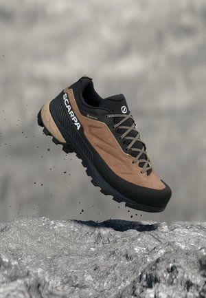 Bruine en zwarte wandelschoen met SCARPA-merk en Gore-Tex-label, weergegeven in de lucht boven rotsachtig terrein met kleine vallende aarde deeltjes.